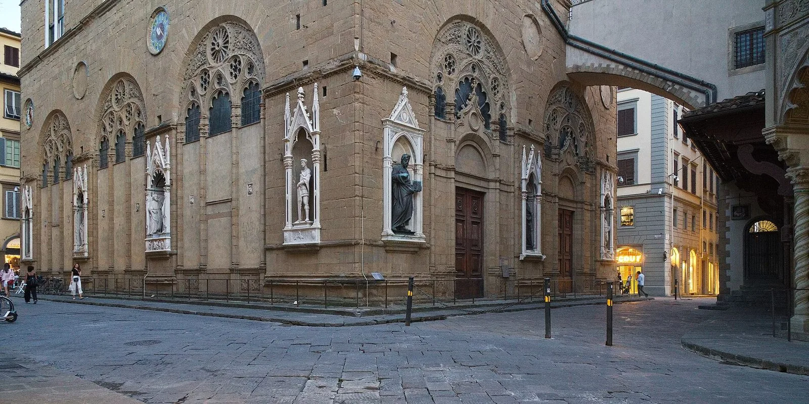 L'angolo della chiesa di Orsanmichele a Firenze: pareti in pietra con grandi archi gotici traforati e nicchie in marmo bianco che ospitano statue di santi, un arco sospeso sulla strada lastricata e alcuni passanti