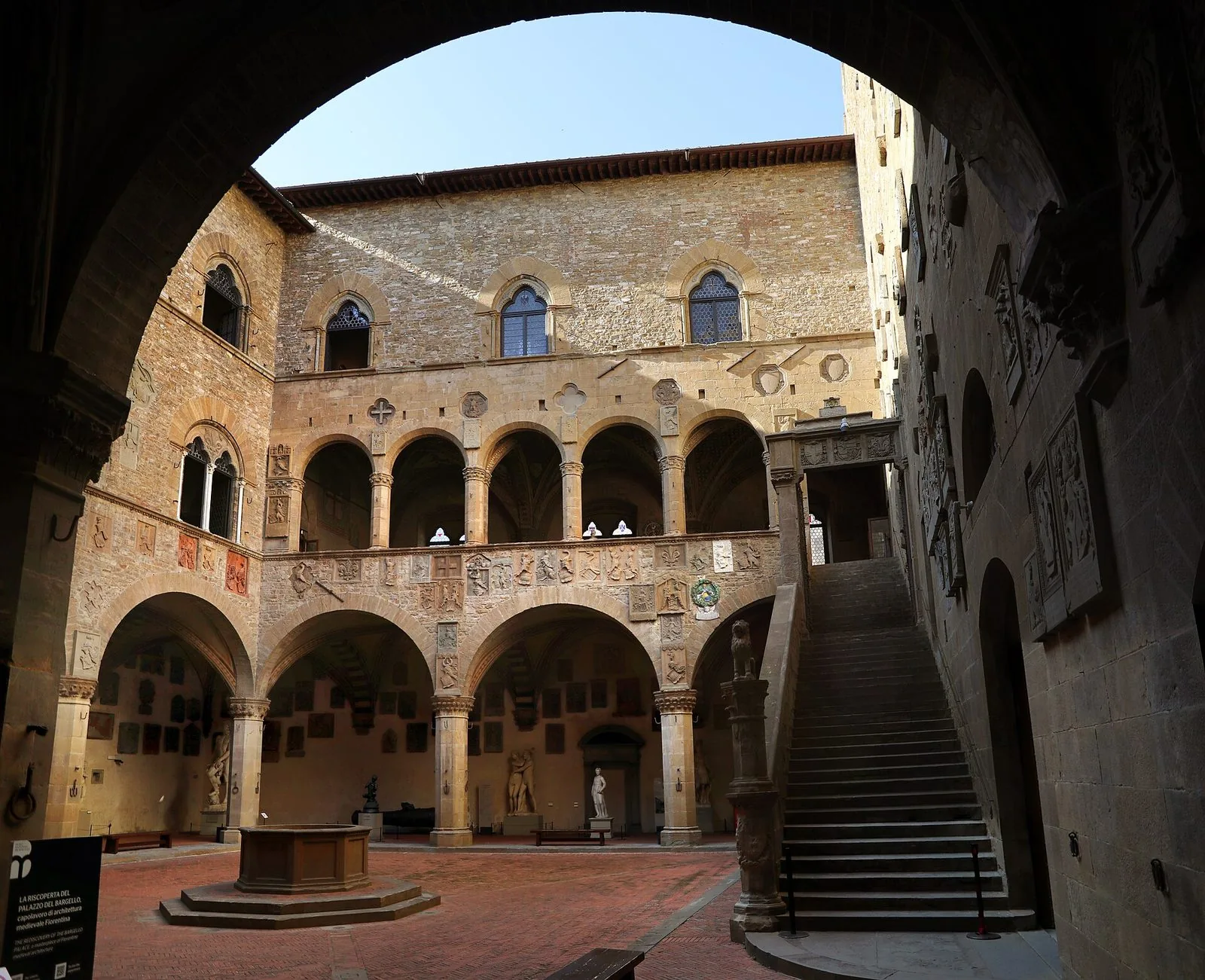 Il cortile del Bargello a Firenze visto da sotto un arco in ombra: porticato con colonne in pietra, loggia al primo piano, pareti ricoperte di stemmi scolpiti, un pozzo ottagonale al centro e una scala esterna in pietra sulla destra